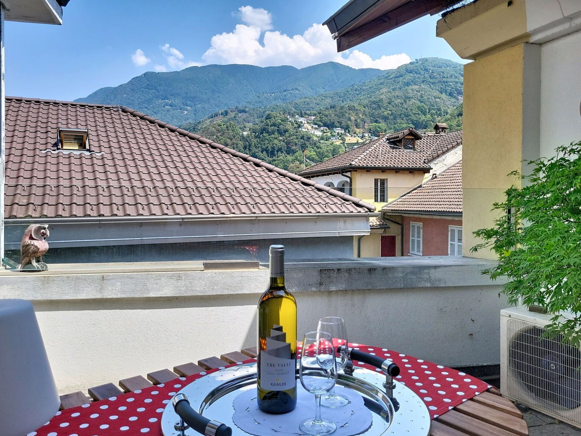 A table with a bottle of wine and glasses, set on a balcony with a view of houses and mountains.