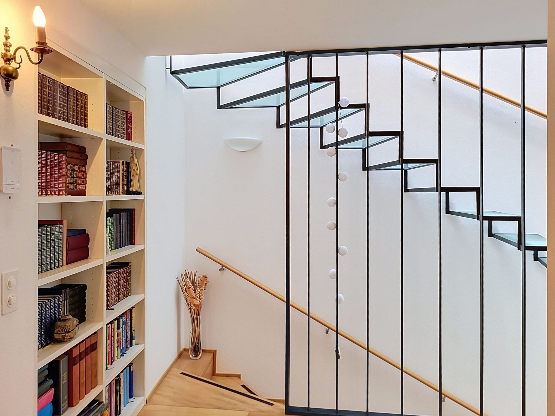 Eine Treppe mit einem geometrischen Geländer, ein Bücherregal voller Bücher und eine dekorative Vase auf dem Boden.
