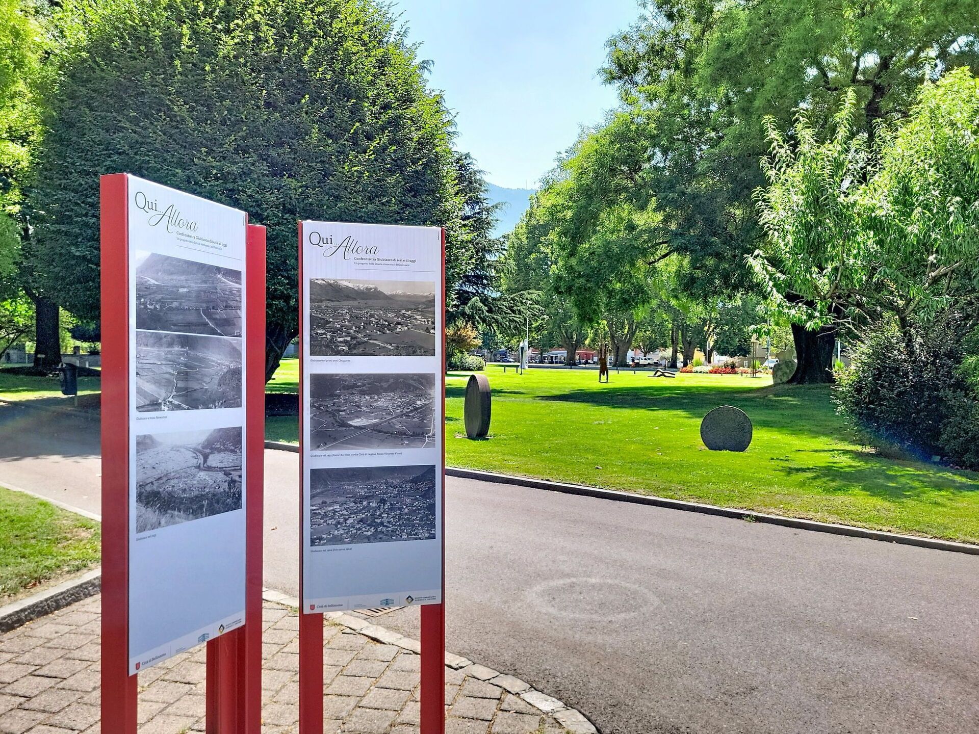Zwei Informationsschilder mit historischen Fotos in einer Freifläche, umgeben von Bäumen und mit einem gepflasterten Weg.