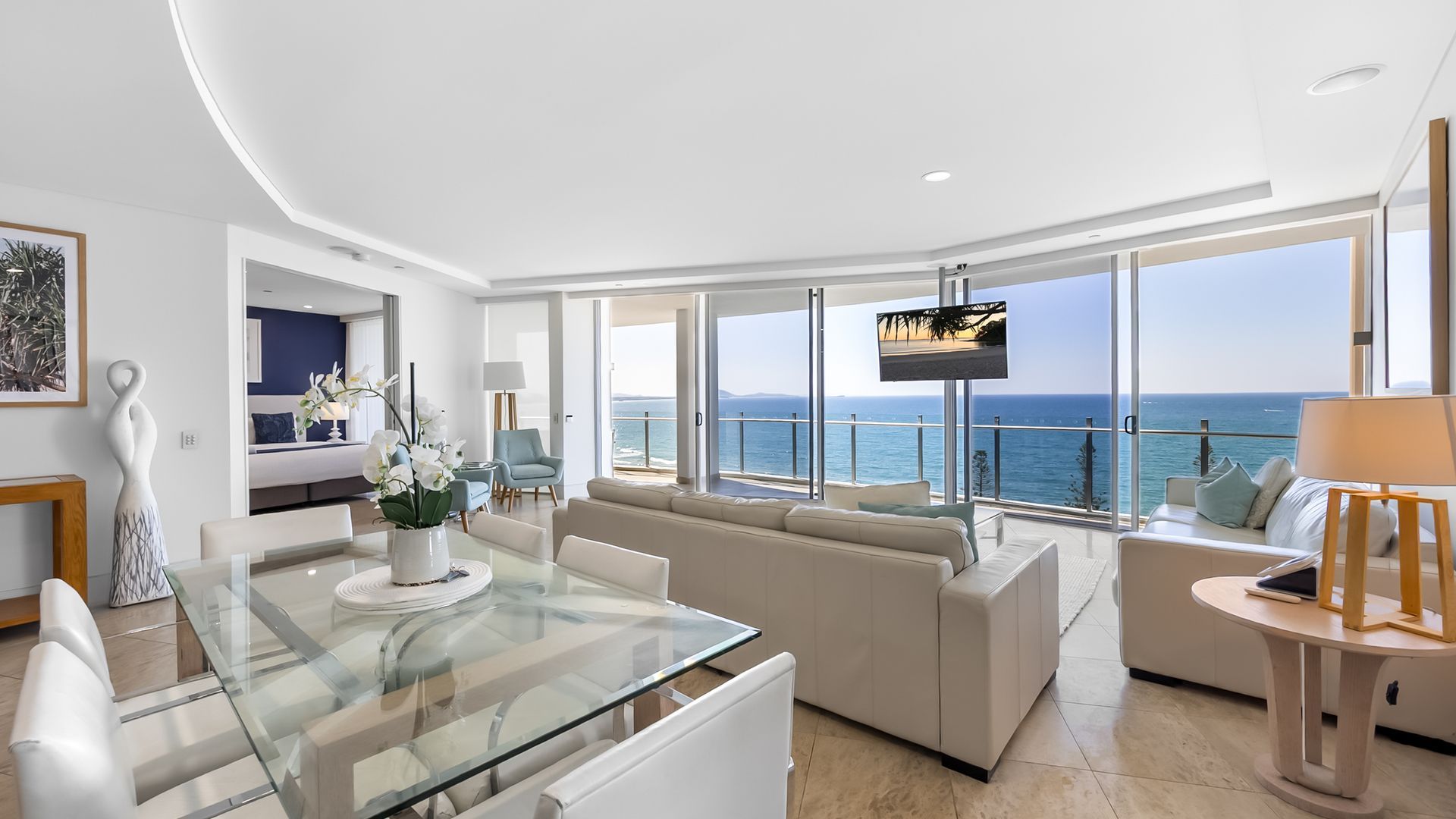 A modern living room with a glass dining table, a sofa, and a TV mounted on the wall.
