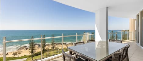 A balcony with a glass table and wicker chairs overlooking a beach and ocean.