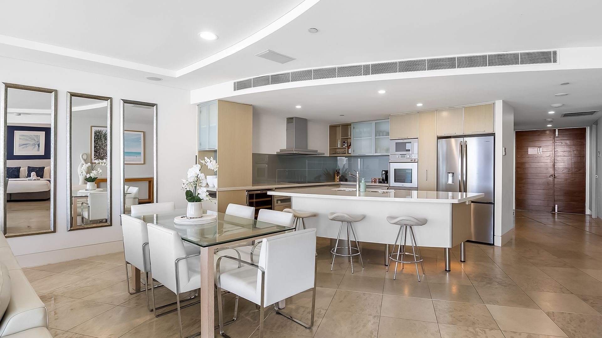 Uma cozinha moderna com ilha central, eletrodomésticos de aço inoxidável e área de jantar com mesa de vidro e cadeiras brancas.