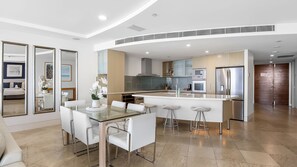 Une cuisine moderne dotée d’un îlot central, d’appareils électroménagers en acier inoxydable et d’un coin repas avec une table en verre et des chaises blanches.