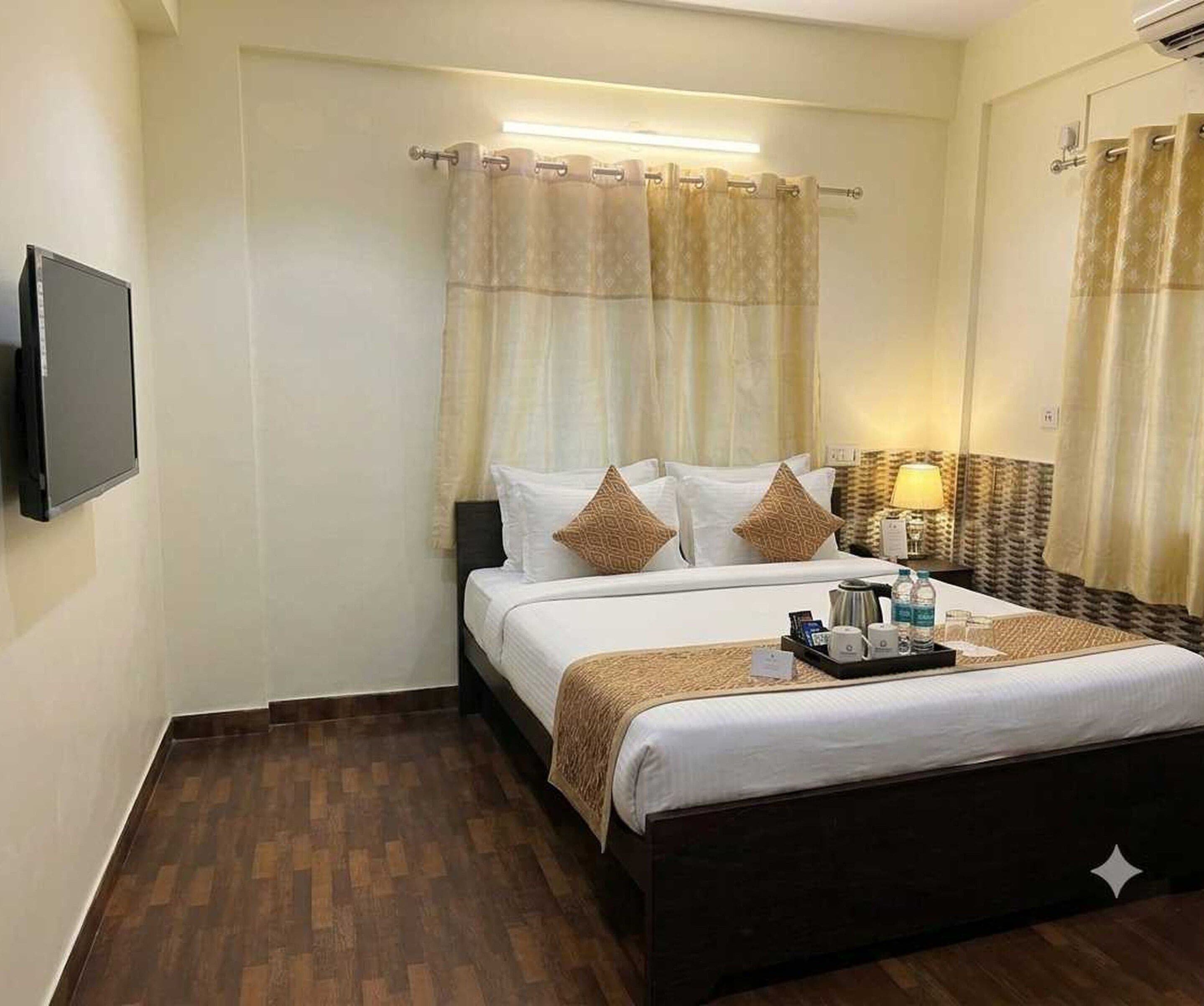 A hotel room with a bed, a bedside table with a lamp, a flat-screen TV mounted on the wall, and a wooden floor.