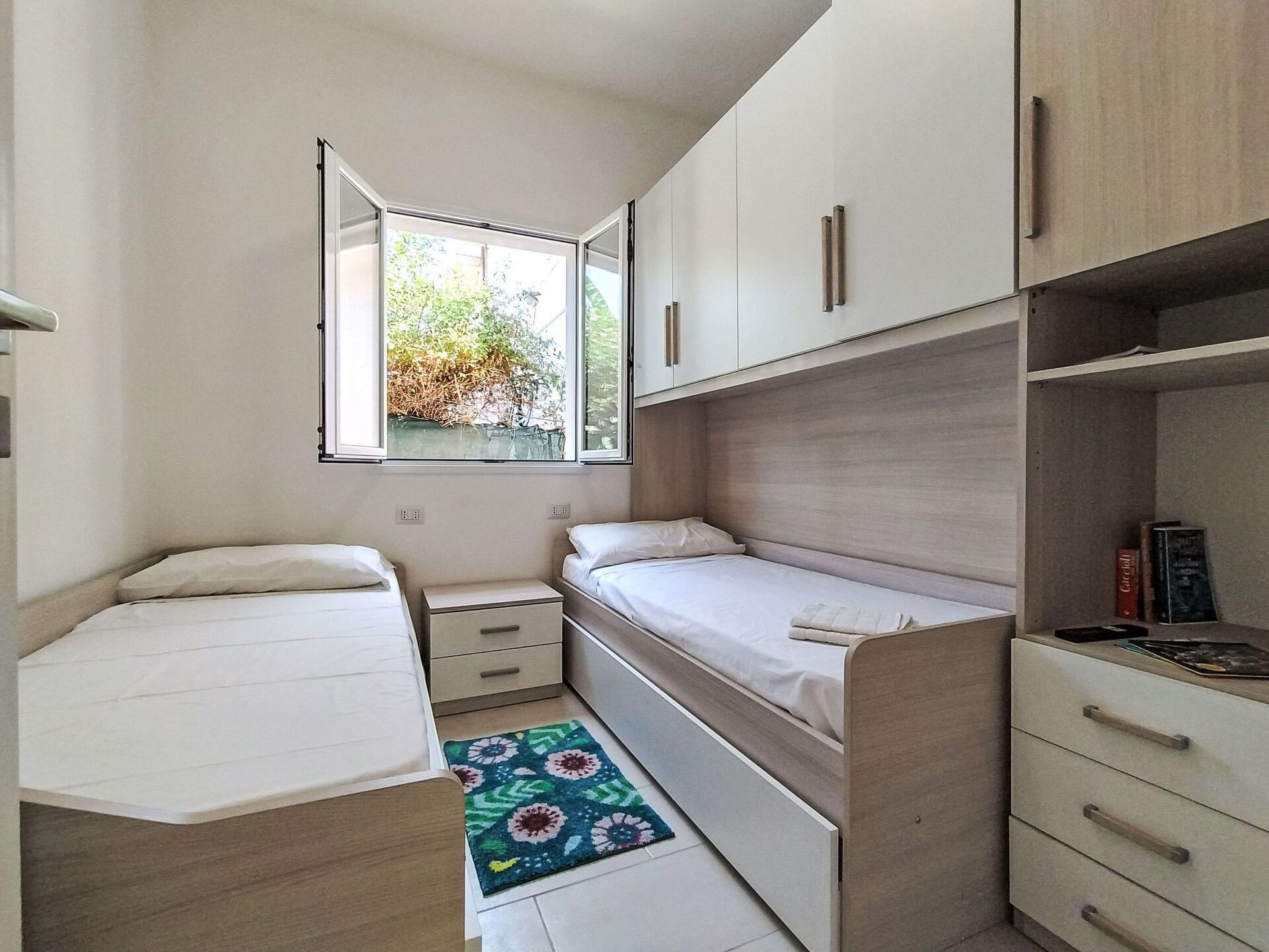 Un dormitorio pequeño con dos camas individuales, una mesita de noche, armario empotrado y ventana con vistas a zonas verdes.