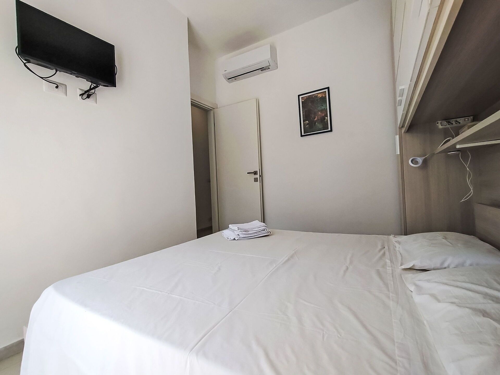 A bedroom with a bed, a flat-screen TV mounted on the wall, an air conditioning unit, and a door leading to another room.