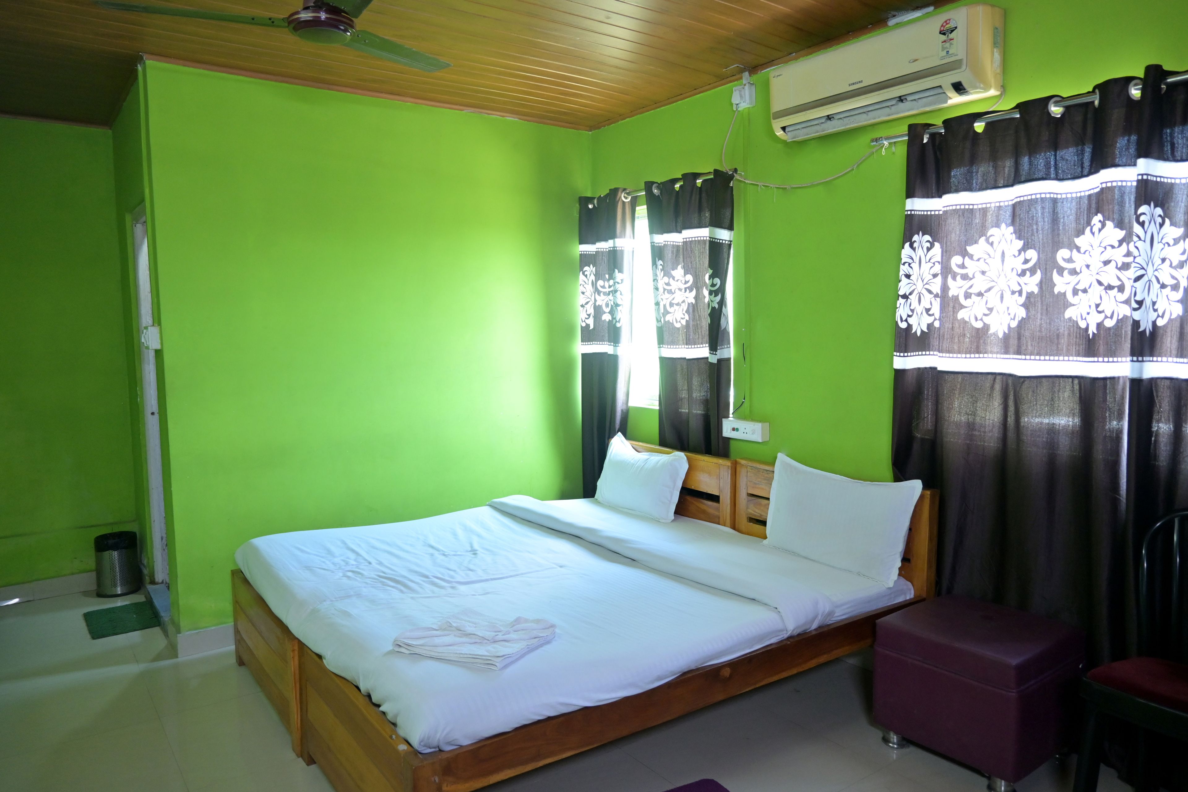 Una habitación con cama, cabecero de madera, ventana con cortinas, unidad de aire acondicionado y una mesita.