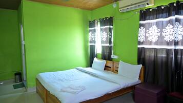 Una habitación con cama, cabecero de madera, ventana con cortinas, unidad de aire acondicionado y una mesita.