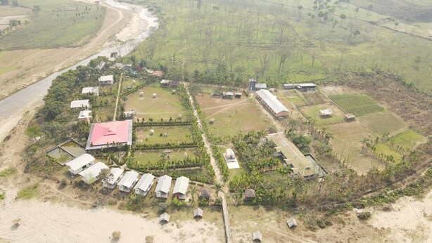 An aerial view of a rural area with a cluster of buildings, some with flat roofs, surrounded by fields and a river. - Khamar Bari (Mal)
