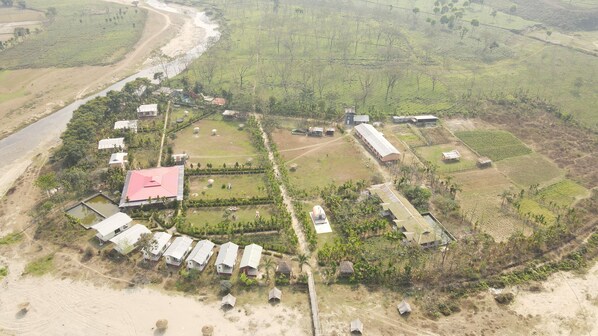 An aerial view of a rural area with a cluster of buildings, some with flat roofs, surrounded by fields and a river.