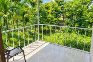 A balcony with a metal railing, a chair, and a view of lush greenery and trees. - Khamar Bari (Mal)
