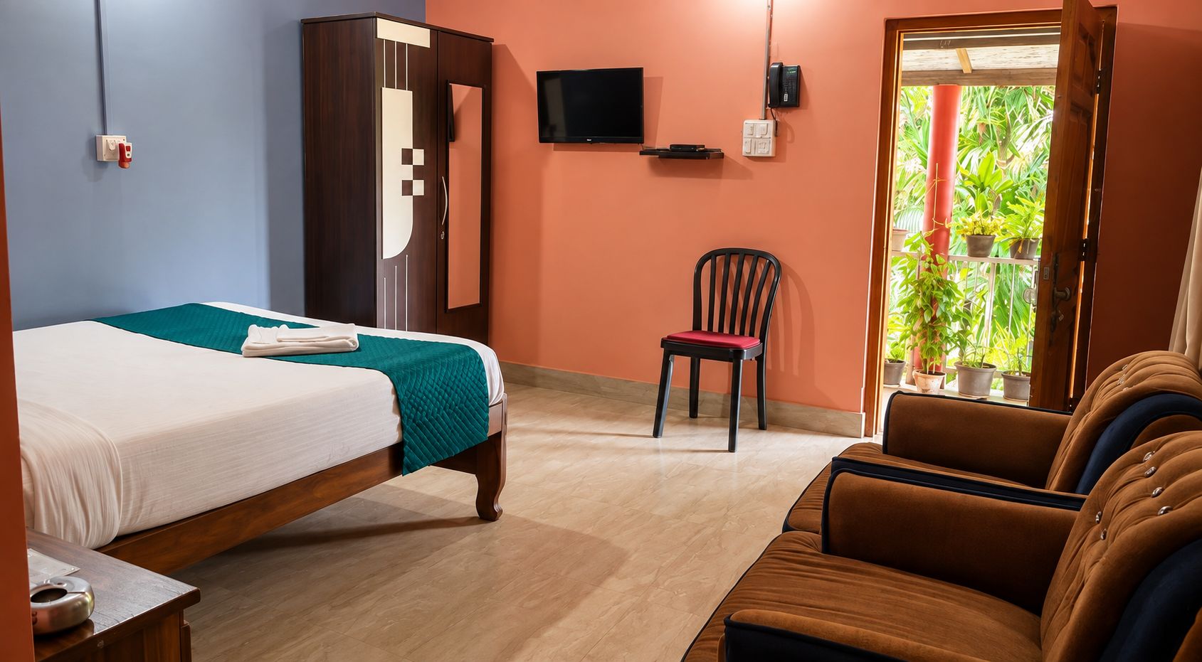 A hotel room with a bed, a chair, a sofa, a television, and a window with plants.