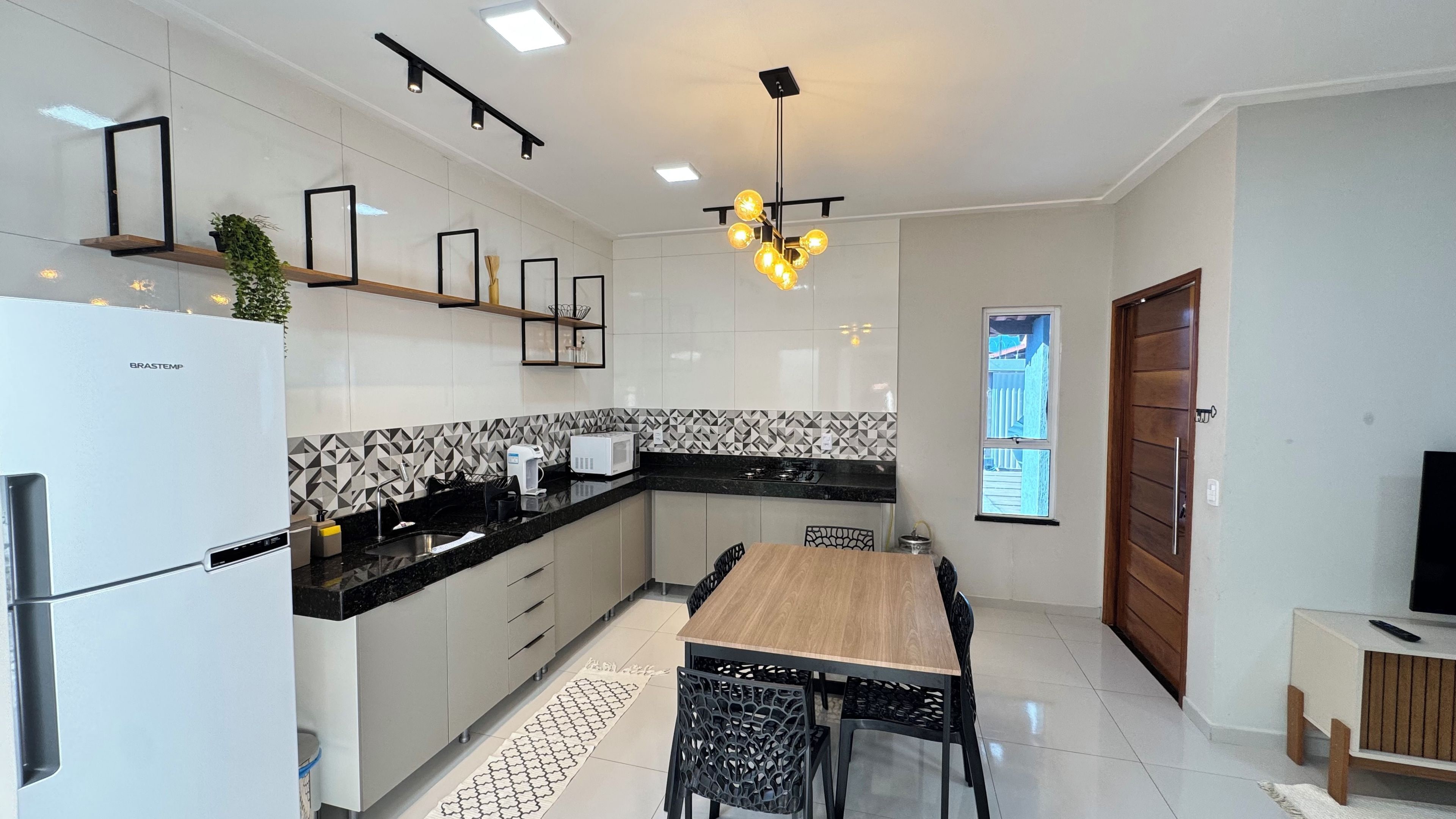 Cozinha moderna com geladeira, micro-ondas e fogão. Há uma mesa de jantar com cadeiras e uma luminária pendente.