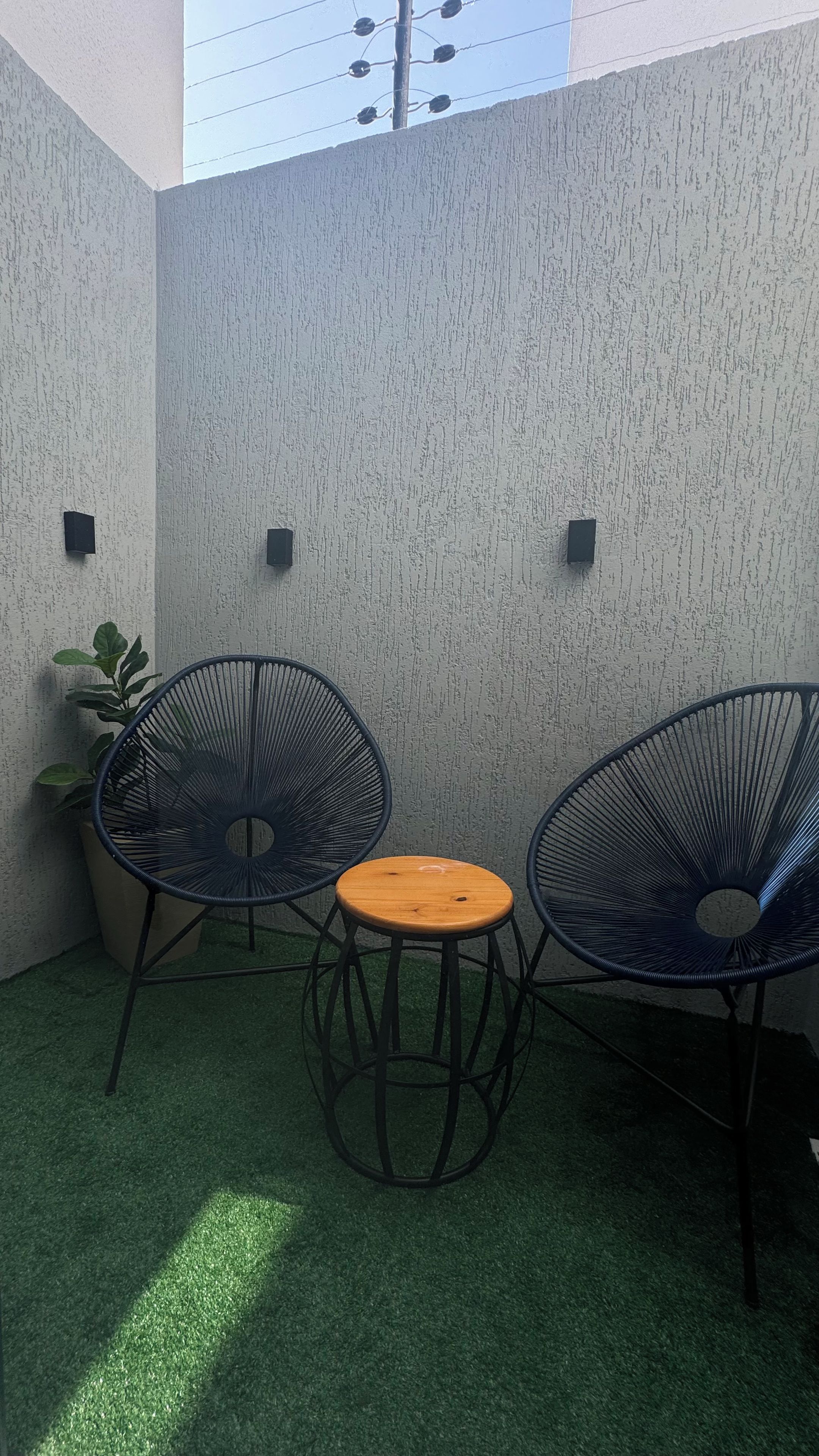Uma pequena área externa com assentos para dois, incluindo cadeiras e uma mesa, cercada por um muro texturizado e um vaso de planta.