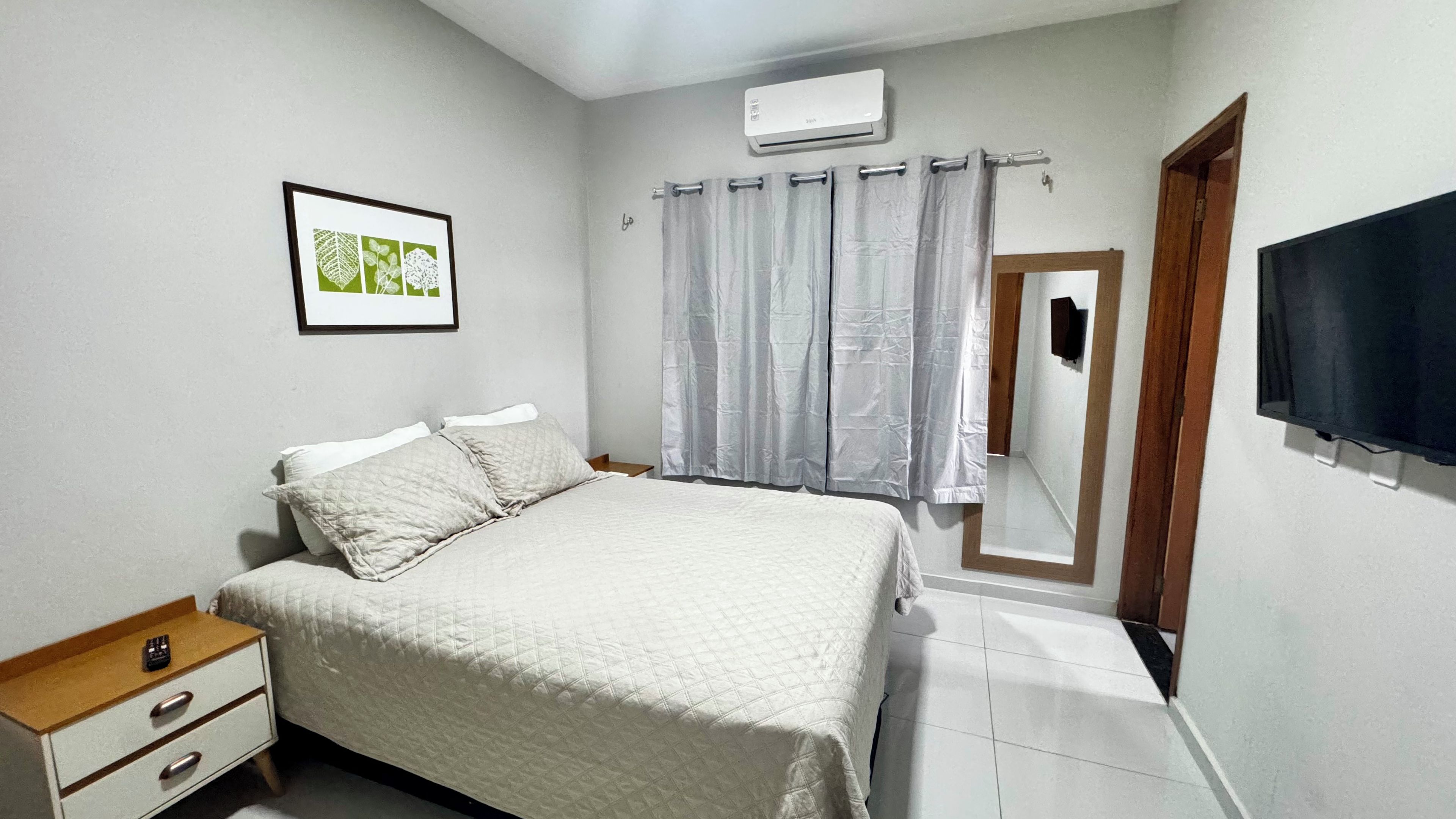 Quarto com cama, mesa de cabeceira, gravura botânica emoldurada, janela com cortinas, ar-condicionado e televisão.
