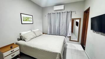 Quarto com cama, mesa de cabeceira, gravura botânica emoldurada, janela com cortinas, ar-condicionado e televisão.