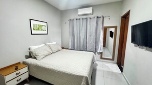 Quarto com cama, mesa de cabeceira, gravura botânica emoldurada, janela com cortinas, ar-condicionado e televisão.