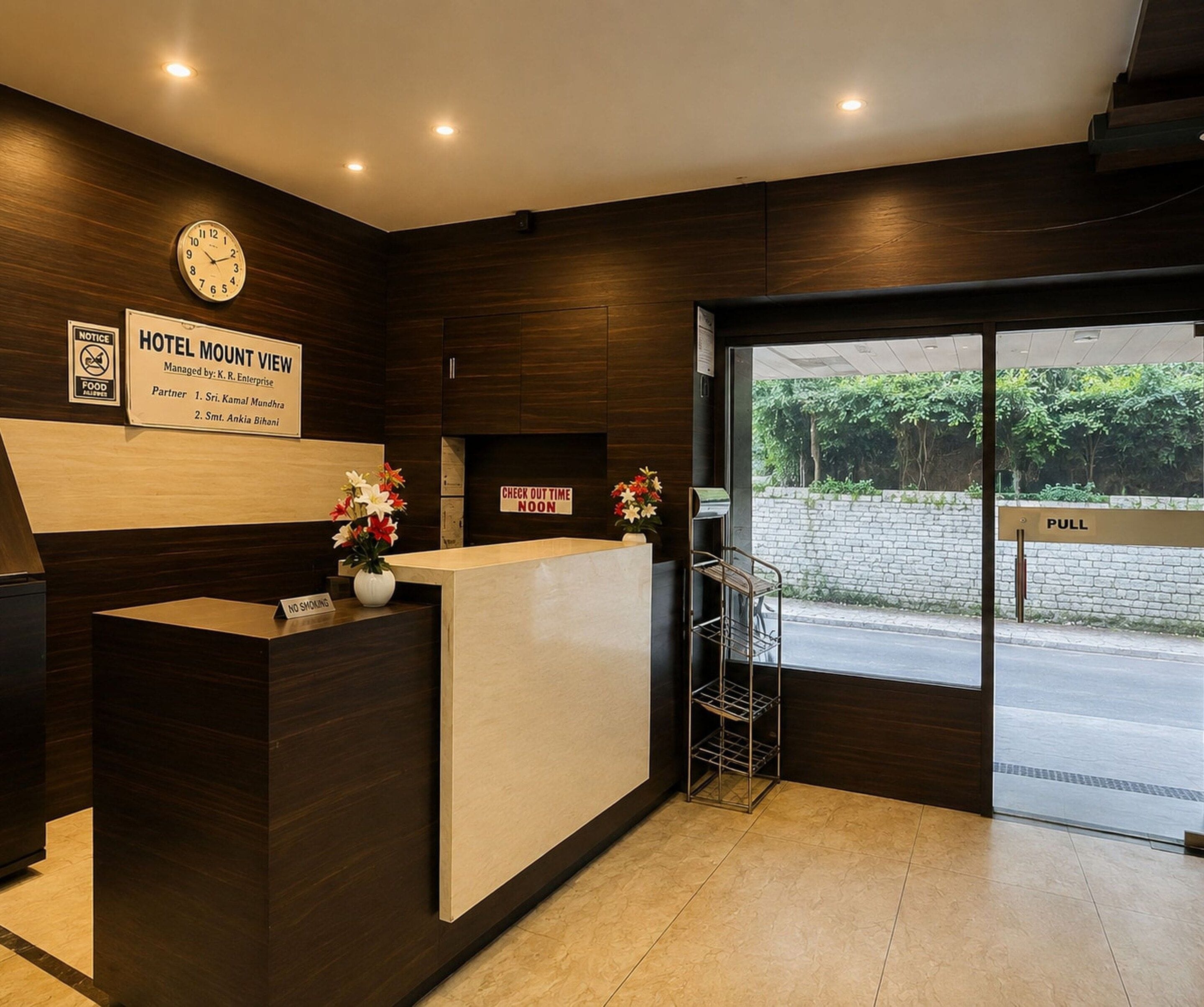 A hotel reception area with a clock, a signboard for Hotel Mount View, and a 'Check Out Time Noon' sign.