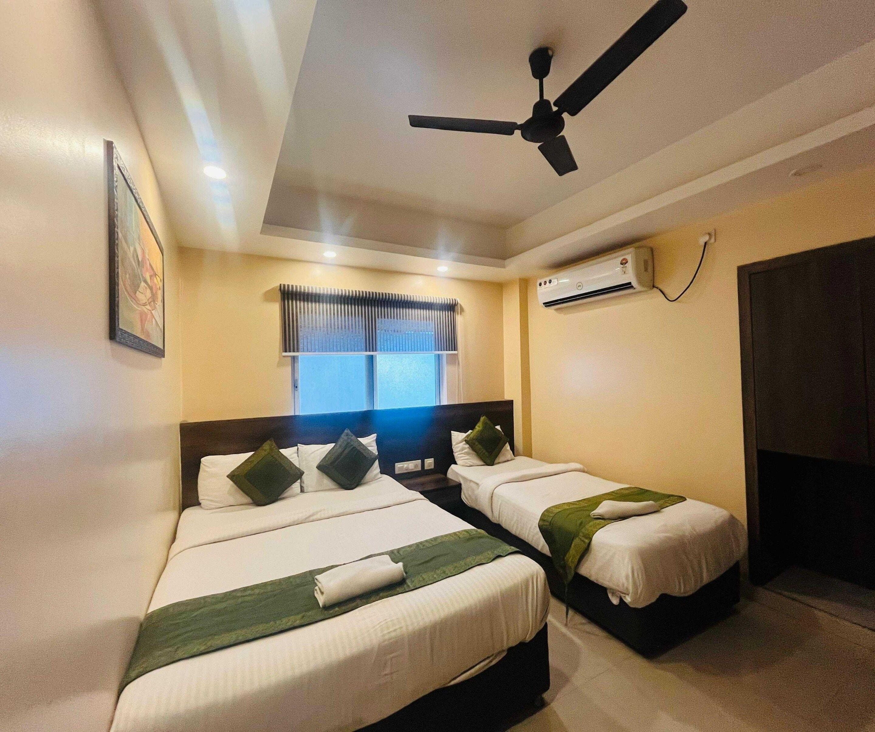 A hotel room with two beds, a ceiling fan, an air conditioning unit, and a window with blinds.