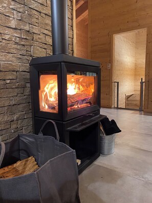 A wood-burning stove with a stone wall backdrop, a metal basket with logs, and a wooden door in the background. - "Serra View" Chalet: Exceptional Views and a Chic, Modern Mountain Atmosphere (Font-Romeu-Odeillo-Via)