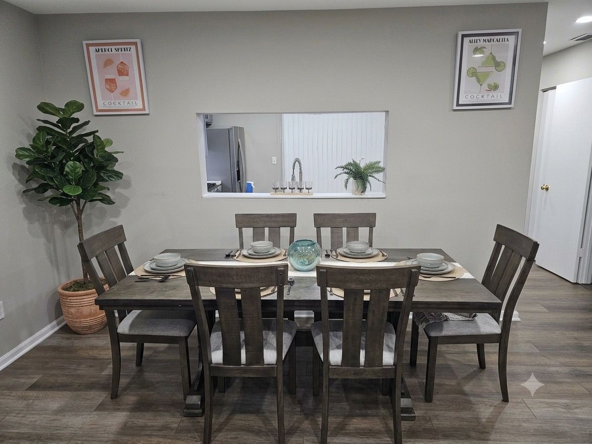 A dining area with a table set for six, surrounded by chairs, a potted plant, and framed artwork on the walls.