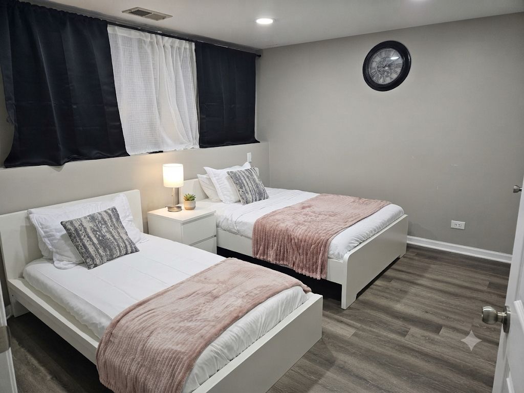 A bedroom with two beds, a bedside table with a lamp, a clock on the wall, and a window with curtains.