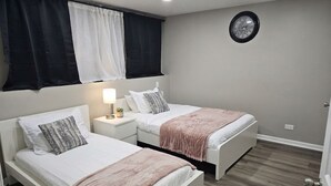 A bedroom with two beds, a bedside table with a lamp, a clock on the wall, and a window with curtains. - 6 Beds Ideal for Crews & Insurance I Close to Naval Base and 6 flags (Waukegan)