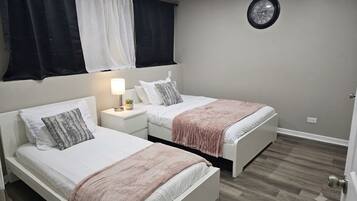 A bedroom with two beds, a bedside table with a lamp, a clock on the wall, and a window with curtains.