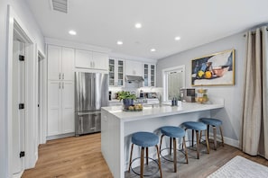 A modern kitchen with white cabinetry, stainless steel appliances, a central island with stools, and a painting above the sink. - Lux Pacific Beach - Rooftop Terrace, Walk to Sand (San Diego)