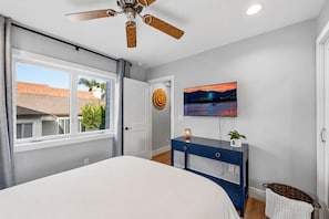 A bedroom with a bed, a ceiling fan, a TV on the wall, and a window with a view of buildings outside. - Lux Pacific Beach - Rooftop Terrace, Walk to Sand (San Diego)