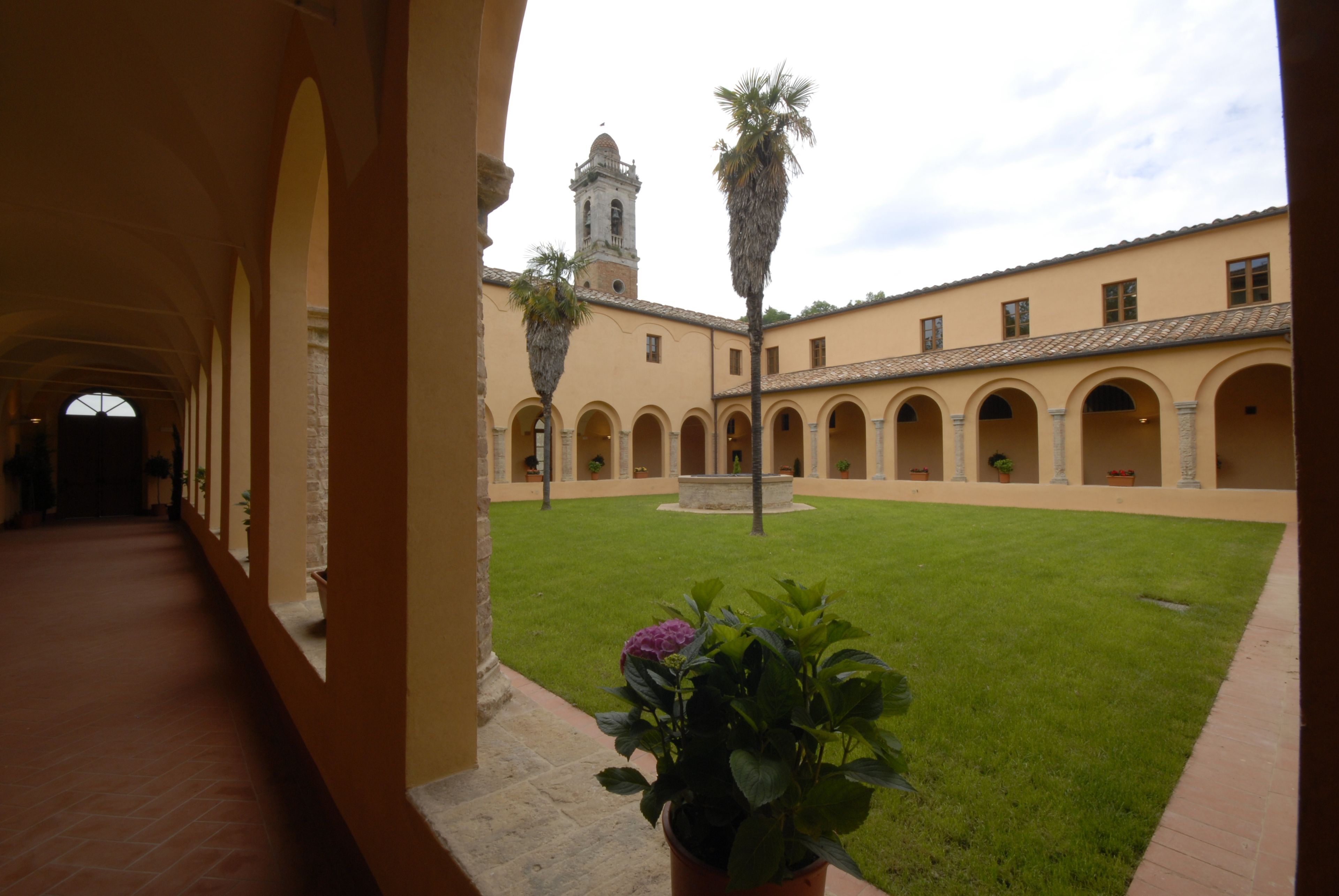 Un cortile con una fontana al centro, circondato da archi e un edificio con una torre sullo sfondo.