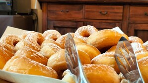 A wooden tray filled with doughnuts dusted with powdered sugar, accompanied by a fork and a knife.