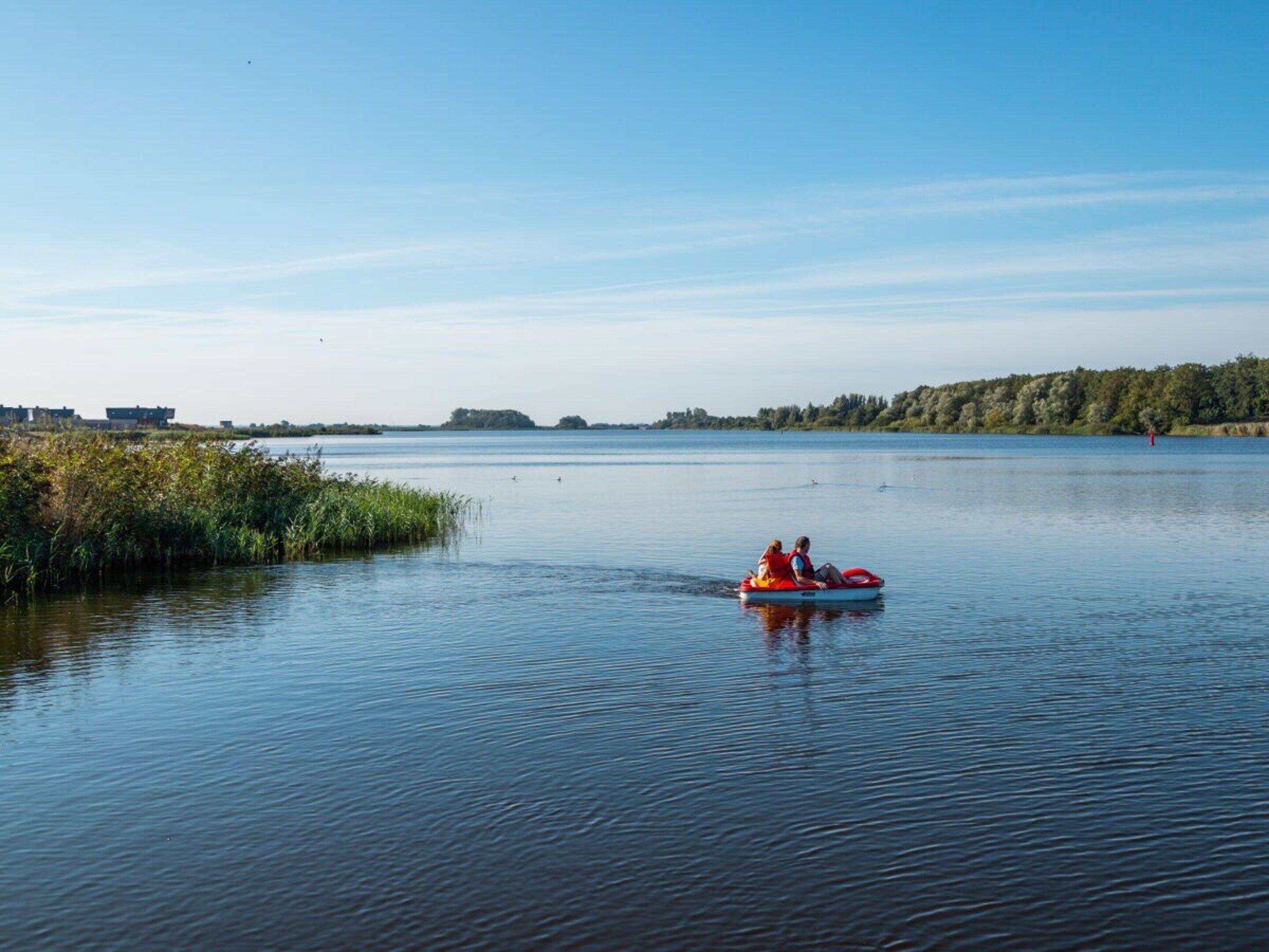A calm lake with a clear sky, a small boat with two people, and a distant shoreline with buildings and trees.
