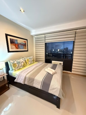A hotel room with a bed, a city view through the window, a framed artwork on the wall, and a bedside table with a lamp. - Stylish Studio with Stunning Makati City Views at Gramercy Residences Makati. (Makati)