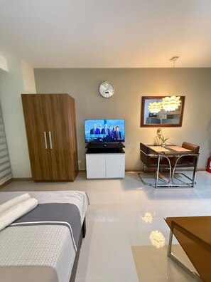 A hotel room with a bed, a desk with a chair, a television, a clock, and a decorative mirror. - Stylish Studio with Stunning Makati City Views at Gramercy Residences Makati. (Makati)