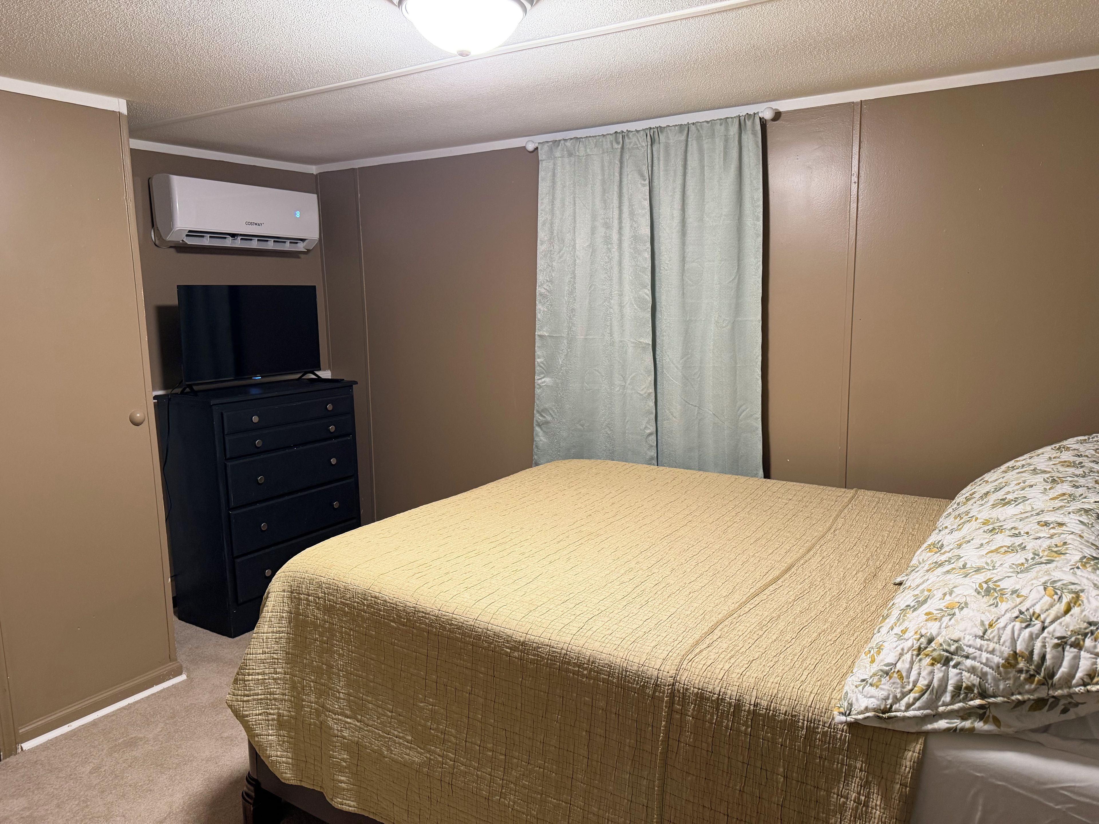 A bedroom with a bed, a dresser, an air conditioning unit, and a window with curtains.