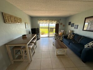 A living room with a sofa, a coffee table, a TV, and a dining area with chairs. - Cozy Ocean & Pool View 2-Bedroom, 2-Bathroom Condo in Amazing Ormond Beach Fla. (Ormond Beach)