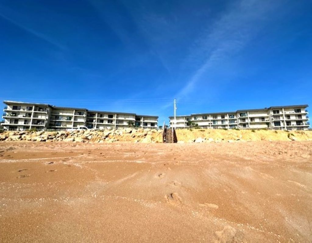 Ein Strandhotel mit mehreren Etagen, ein Sandstrand im Vordergrund und ein klarer, blauer Himmel.