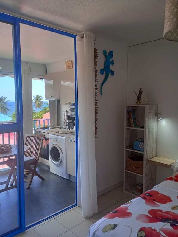 Une pièce comprenant un lit, un lave-linge, une petite table avec des chaises et une vue sur l’océan, visible à travers une porte vitrée.