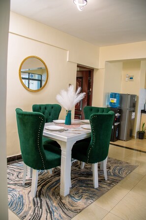 A dining area with a round table, four green upholstered chairs, a white tablecloth, and a decorative vase with white feathers. - Kileleshwa Gem|Luxury 2b near Westlands (Nairobi)