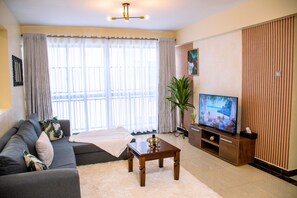 A living room with a sofa, a coffee table, a TV, and a large window with curtains. - Kileleshwa Gem|Luxury 2b near Westlands (Nairobi)