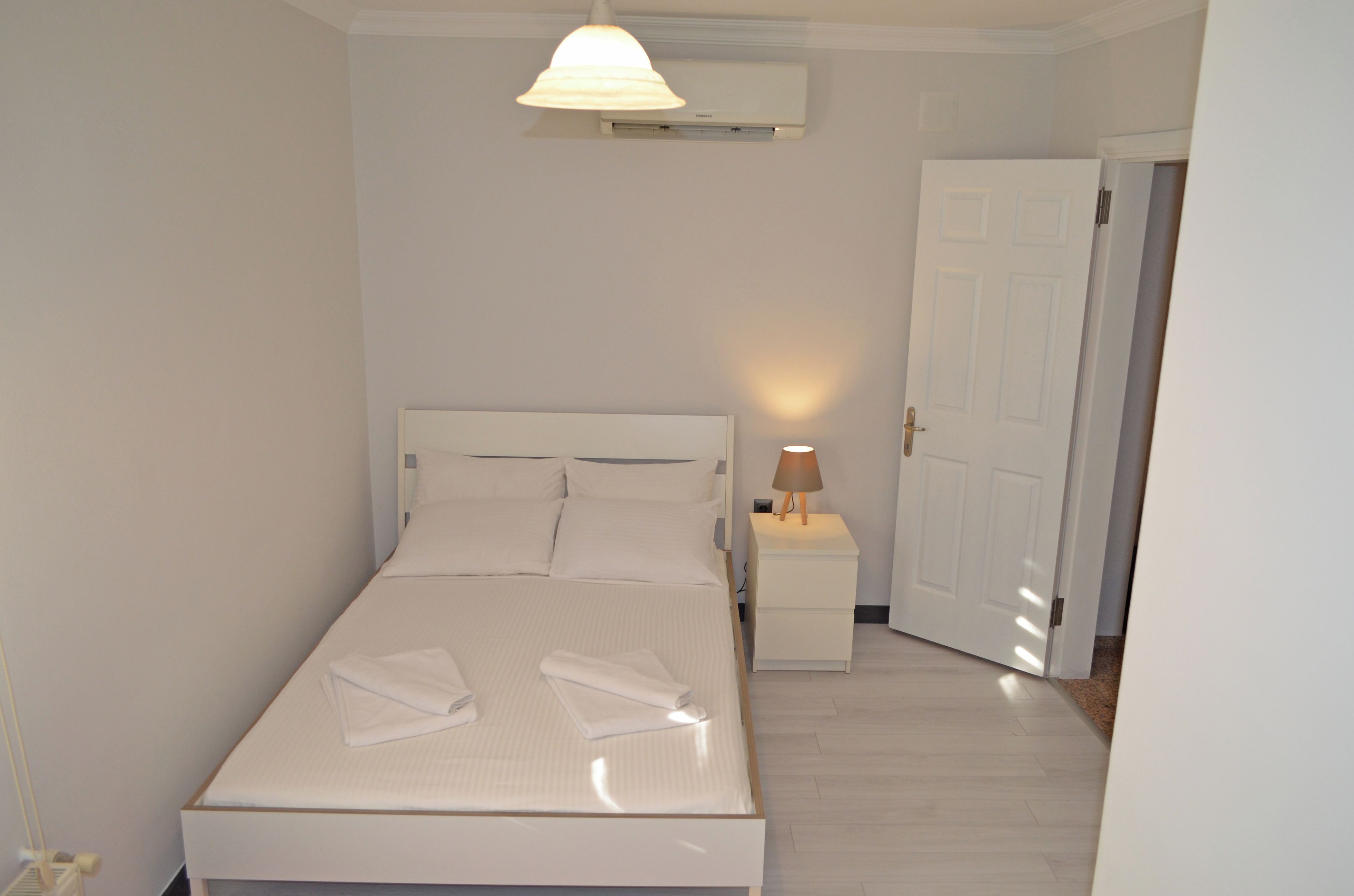 Una cama bien hecha con almohadas y una toalla doblada, una lámpara de noche y una puerta a la derecha.