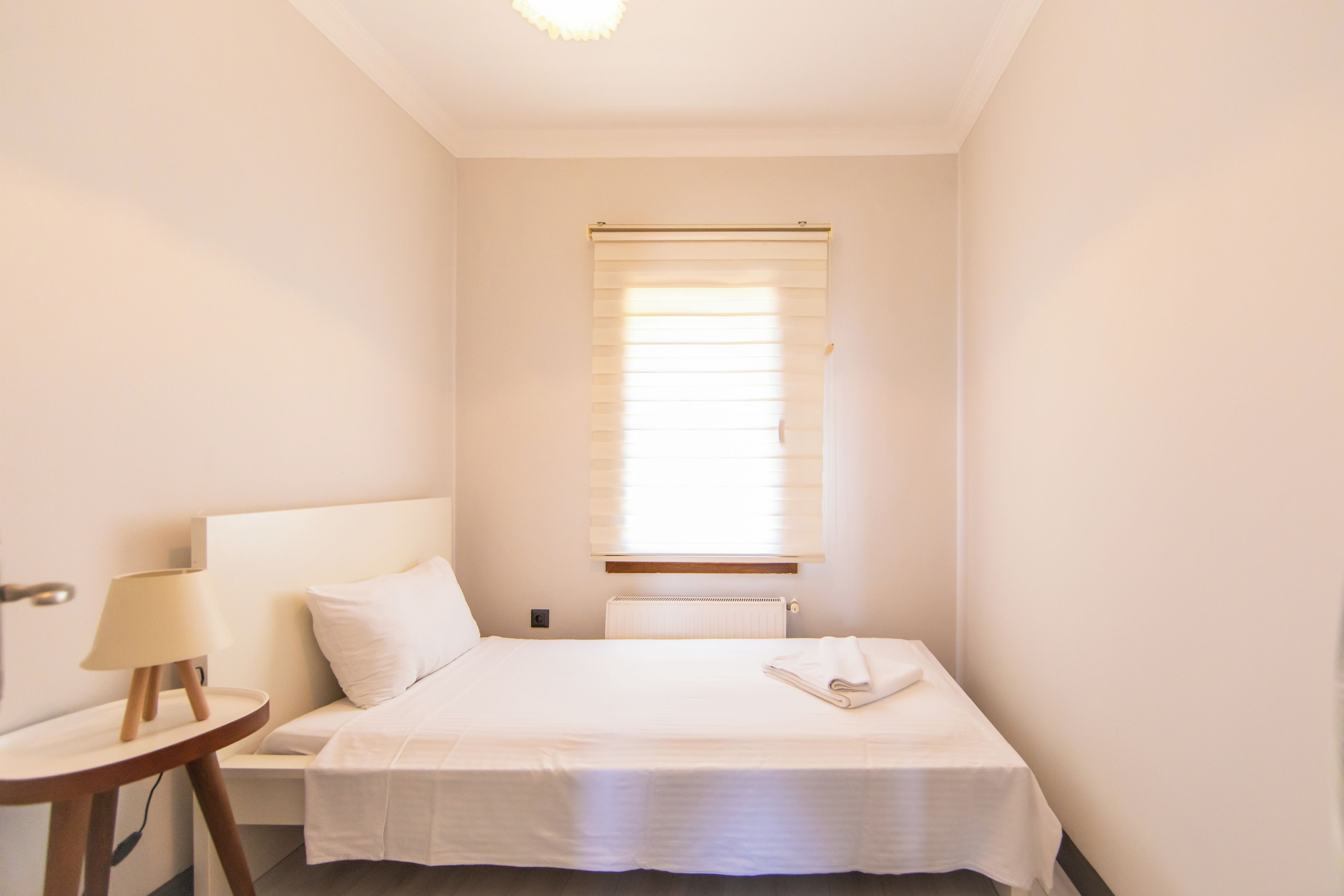 Ein kleines, sauberes Zimmer mit einem Einzelbett, einem Nachttisch mit Lampe und einem Fenster mit Jalousien.