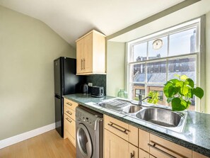 A compact kitchen with a washing machine, a black refrigerator, wooden cabinets, a window with a view of buildings, and a sink. - Cozy 1BR in New Town - Steps from the Royal Mile (Edinburgh)