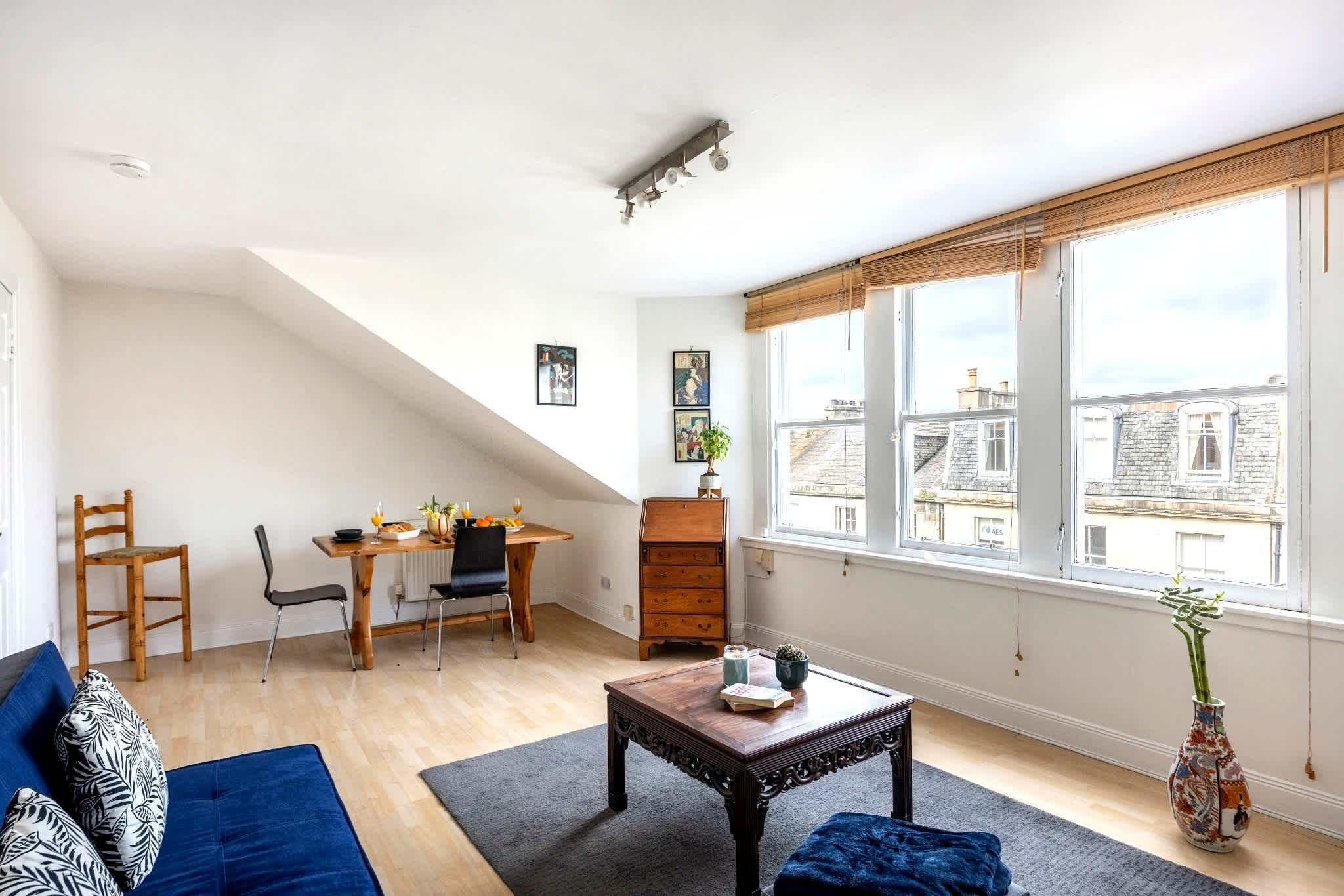 A living room with a sofa, coffee table, dining table, chairs, and a large window with a view of buildings.