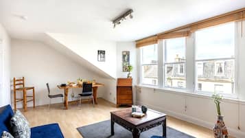 A living room with a sofa, coffee table, dining table, chairs, and a large window with a view of buildings.