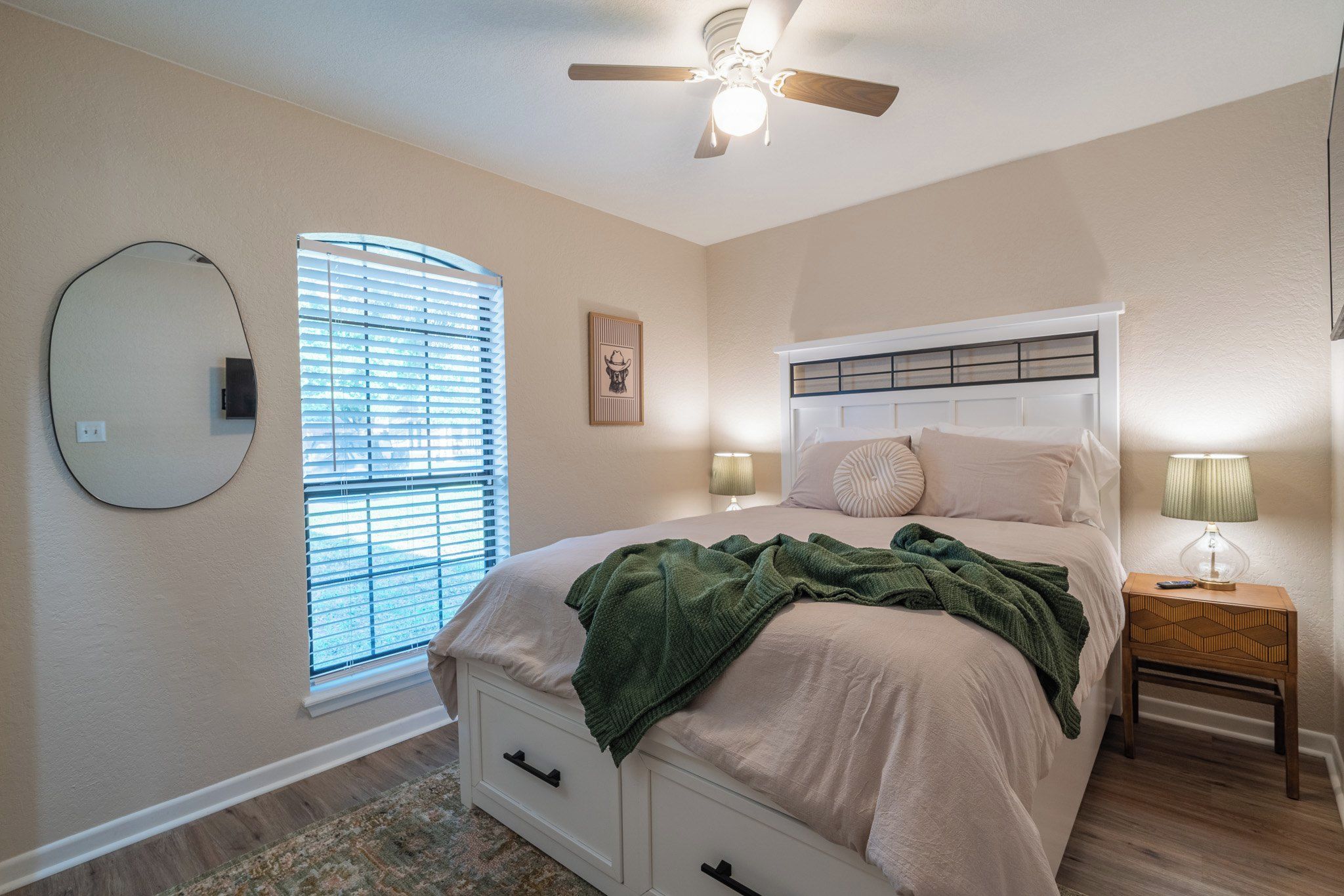A bedroom with a bed, bedside tables, a ceiling fan, a window with blinds, and a wall-mounted mirror.