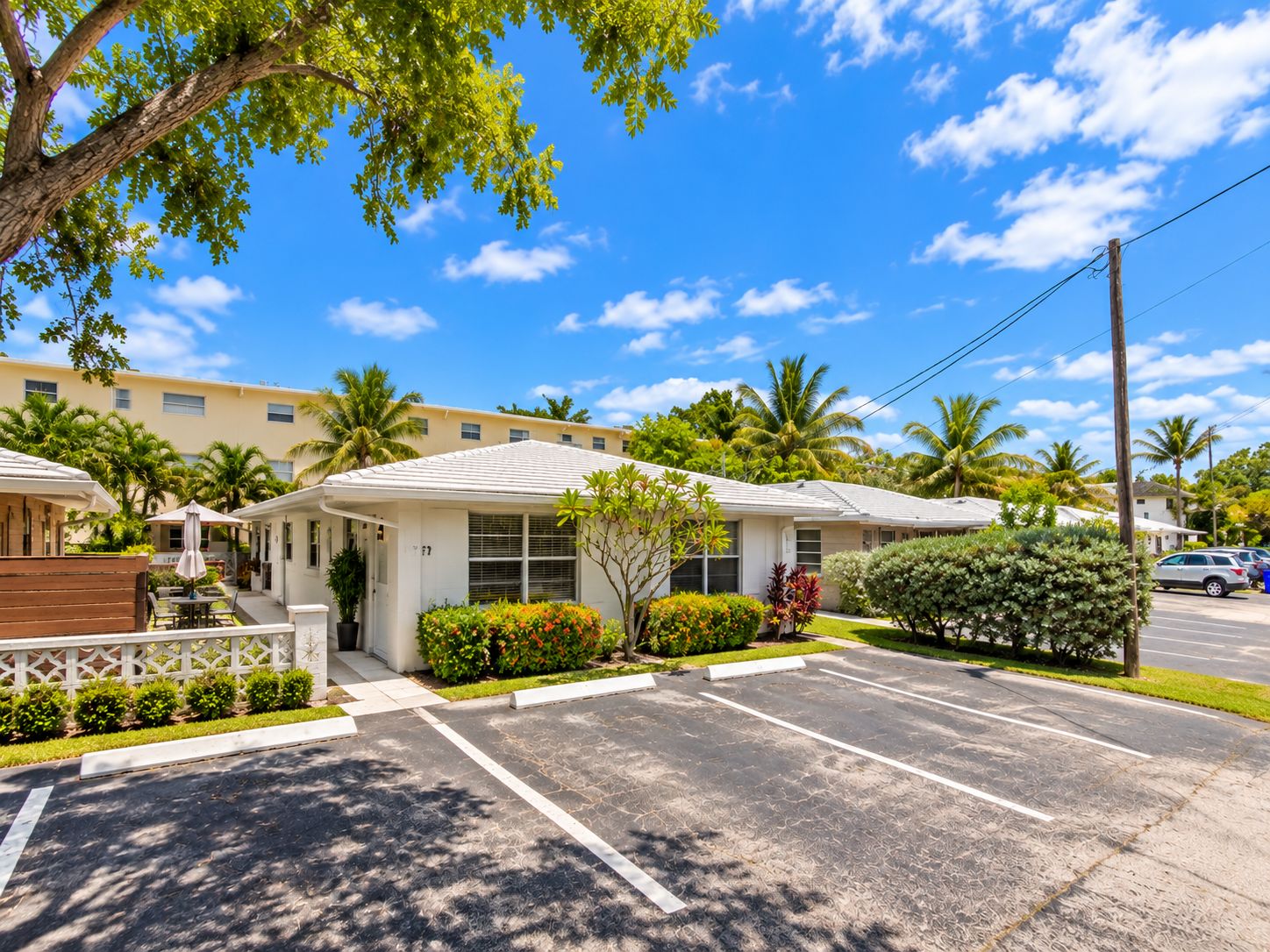 A hotel with a parking lot, a covered outdoor seating area, and a view of palm trees and other greenery.