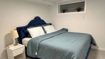 A bedroom with a bed, bedside table, lamp, and a window with plants.