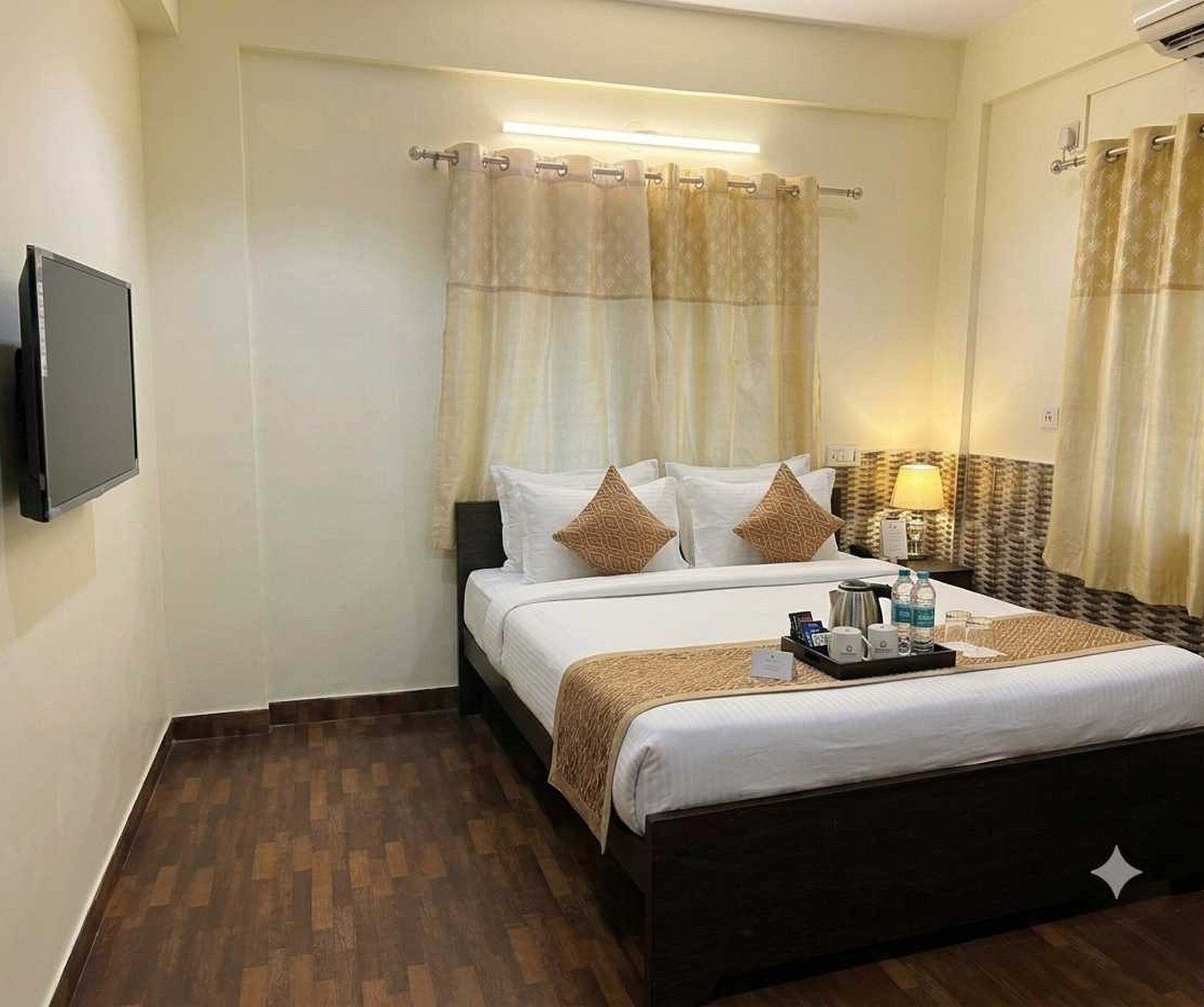 A hotel room with a bed, a bedside table with a lamp, a flat-screen TV mounted on the wall, and a wooden floor.